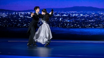 Un bailarín con esmoquin negro y un robot humanoide con un vestido gris y negro realizan un paso de baile sobre un escenario azul. Fondo de ciudad nocturna iluminada