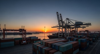 Imagen aérea de un puerto comercial al amanecer con múltiples grúas, barcos de carga y cientos de contenedores de envío apilados en el muelle.