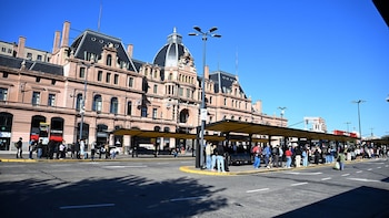 Calle con paradas de autobús amarillas llenas de gente esperando, frente a un gran edificio histórico rosado con cúpula, bajo un cielo azul claro