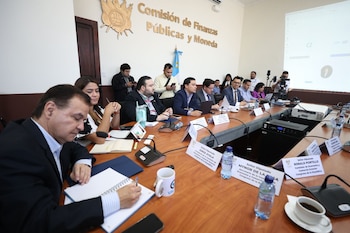 Varias personas, mayormente hombres, sentadas alrededor de una mesa de reuniones de madera, con micrófonos, computadoras portátiles y documentos. Un hombre escribe en un cuaderno