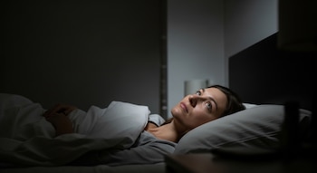 Retrato horizontal de una mujer morena acostada en la cama, cubierta con sábanas blancas, mirando hacia arriba en una habitación oscura.