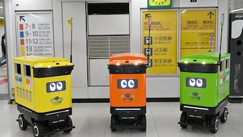 Tres pequeños vehículos robóticos de colores amarillo, naranja y verde con ojos digitales están alineados dentro de una estación de tren