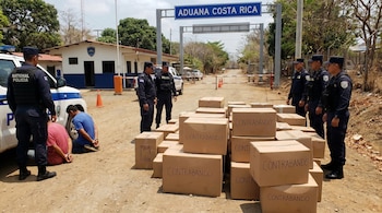 Agentes de la Policía Nacional de Costa Rica custodian a dos personas esposadas junto a pilas de cajas marcadas "Contrabando" en un puesto fronterizo.