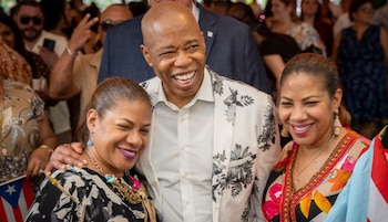 Eric Adams, vestido con un traje blanco y negro floreado, ríe alegremente entre dos mujeres sonrientes en un evento con banderas puertorriqueñas