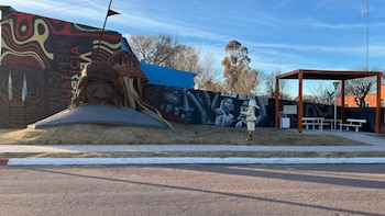 Un gran monumento exterior con una escultura de cabeza, murales de rostros y manos, texto "VERDAD JUSTICIA MEMORIA", y una pérgola de madera con bancos