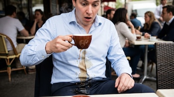 Un hombre con camisa azul derrama café caliente sobre su ropa, creando una gran mancha marrón. Su expresión es de asombro mientras sostiene la taza.