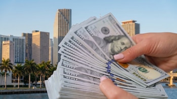 Primer plano de una mano contando billetes de cien dólares estadounidenses con el horizonte de Miami, edificios y palmeras bajo un cielo azul despejado al fondo.