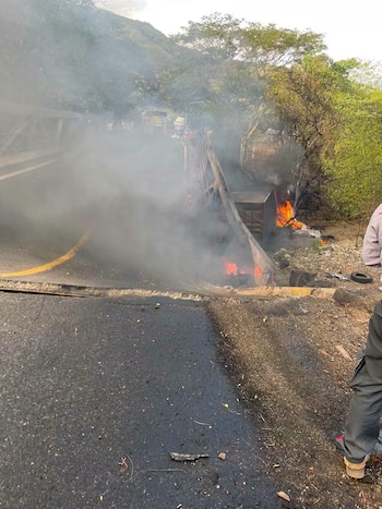 Carretera con asfalto dañado, humo denso, llamas en el barranco junto a la estructura de un puente y árboles al fondo
