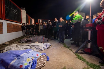 Personas de pie y agachadas durante la noche, tres cuerpos cubiertos con mantas en el suelo, oficial de policía, luz artificial brillante