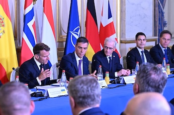 Hombres con traje sentados en una mesa con mantel azul frente a banderas de España, Noruega, Hungría, OTAN, Alemania, Reino Unido, botellas de agua y micrófonos