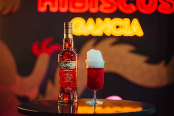 Una botella de licor rojo y una copa con líquido rojo cubierta de espuma sobre una mesa oscura frente a un letrero de neón