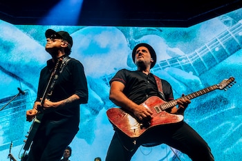Andrés Calamaro tocando la guitarra eléctrica con un sombrero en un escenario iluminado de azul durante un concierto, junto a otro guitarrista