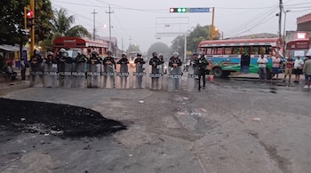 Diez policías con uniformes, cascos y escudos transparentes con la palabra "POLICIA" bloquean una calle. Se observan dos buses de transporte público y personas