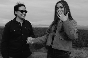 Fotografía en blanco y negro de un hombre con gafas de sol y una mujer que enseña un anillo en su mano izquierda frente a un campo con árboles
