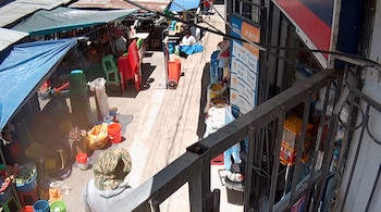 Vista superior de un pasaje comercial con toldos, sillas de plástico apiladas, baldes de colores y una baranda metálica en primer plano.