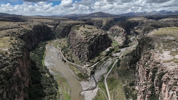 Vista aérea de un cañón rocoso con un río marrón serpenteando. Grandes formaciones rocosas cubiertas de vegetación árida y caminos de tierra, bajo un cielo nublado