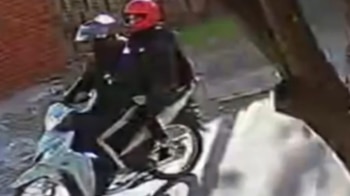 Dos personas con cascos, uno rojo y otro blanco, montan una motocicleta en una calle urbana con un edificio de ladrillos y árboles