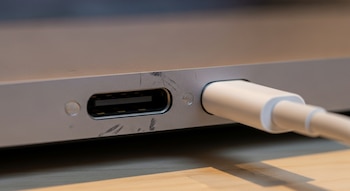 Puerto USB-C de un MacBook con un cable de carga conectado, sobre una superficie de madera clara.