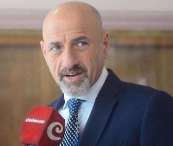 Primer plano de hombre calvo con barba y bigote, vestido con traje oscuro y corbata a puntos, hablando frente a un micrófono rojo de Crónica
