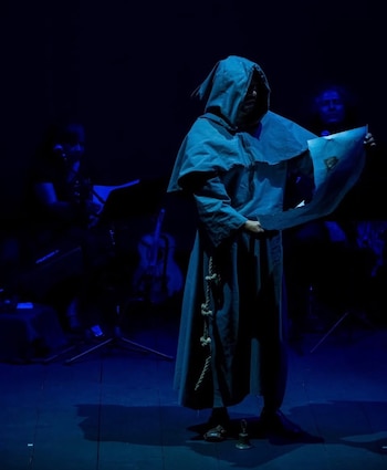 Una persona vestida con túnica y capucha lee un papel en un escenario iluminado con luz azul, junto a músicos al fondo