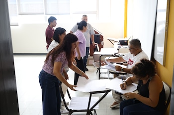 Un grupo de personas en un aula, algunas sentadas y otras de pie, organizan y revisan documentos electorales sobre escritorios escolares