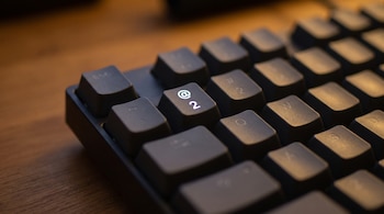 Primer plano de un teclado de computadora de color oscuro con una tecla que muestra el signo arroba y el número dos iluminada con luz morada.