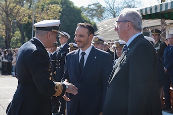 Hombre en uniforme naval saluda a un hombre en traje azul oscuro. A la derecha, otro hombre en traje oscuro y gafas. Fondo con personas y árboles