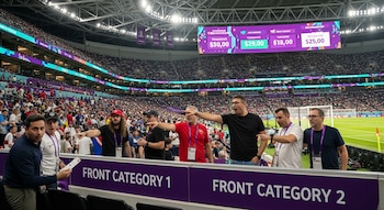 Vista interior de un estadio de fútbol con un campo verde, portería y asientos llenos de público. Pantallas digitales muestran precios; varias personas observan y señalan.