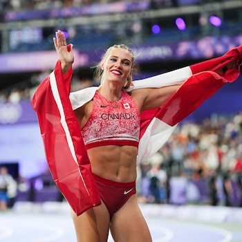 Alysha Newman, atleta rubia, sonríe y saluda, envuelta en la bandera canadiense. Viste un top deportivo rojo y blanco con "CANADA". Fondo borroso del estadio