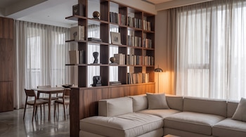 Sala de estar con sofá esquinero claro, estantería de madera oscura con libros y adornos, mesa de comedor con sillas y cortinas blancas en ventanas grandes.