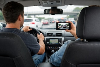 Interior de un auto visto desde atrás. Un conductor sujeta el volante, mientras un pasajero en el asiento delantero derecho graba el tráfico con un celular.