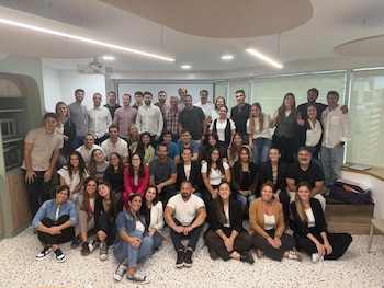 Un grupo diverso de aproximadamente cuarenta personas posa sonriendo en una sala, con hombres y mujeres de diferentes edades vestidos casualmente