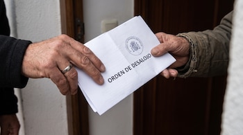 Dos manos sostienen un documento de papel blanco con el texto impreso orden de desalojo y un sello judicial frente a una puerta.