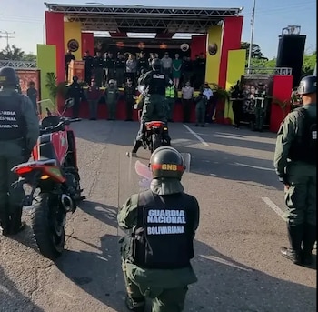 Integrantes de la Guardia Nacional Bolivariana portan escudos y chalecos junto a motocicletas frente a un escenario decorado en rojo y amarillo