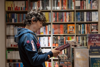 Bruno Pinasco, con cabello rizado y una sudadera azul, sostiene un libro rojo mientras mira las estanterías repletas de volúmenes en una librería