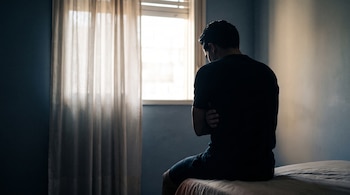 Silueta de un hombre sentado de espaldas en el borde de una cama con los brazos cruzados mirando hacia una ventana iluminada.