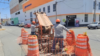 Dos operarios con casco naranja trabajan con maquinaria en una calle delimitada por conos y mallas de seguridad naranjas
