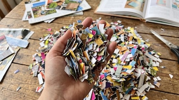Una mano sostiene pequeños trozos de papel recortado sobre una mesa de madera con tijeras, hojas impresas y un libro abierto.