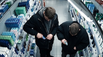 Vista cenital de dos hombres en un pasillo de supermercado guardando envases de productos en los bolsillos de sus abrigos.
