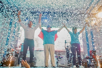 Tres personas en un escenario, el hombre central con camiseta azul y capa roja, flanqueado por un hombre y una mujer, todos con brazos alzados y confeti cayendo