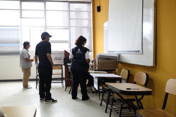 Tres personas en una sala con ventanas grandes y una pared amarilla, preparando una mesa electoral con una urna y cajas de la ONPE, junto a escritorios y sillas