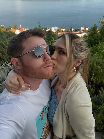 Primer plano de una mujer rubia con pendientes de aro dorados besando la mejilla de un hombre barbudo con gafas de sol, con casas y mar al fondo