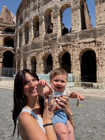 Ailén Cova, con cabello oscuro, sonríe sosteniendo a una bebé con pañal azul y camiseta blanca, frente al Coliseo de Roma bajo un cielo azul