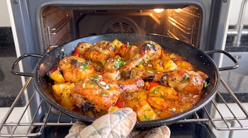 Una mano con guante de cocina retira de un horno abierto una paellera negra con pollo asado, patatas y pimientos.