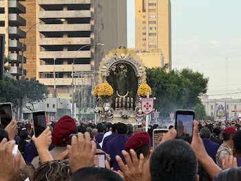 Una multitud observa y fotografía con teléfonos móviles un anda religiosa dorada que avanza por una avenida flanqueada por edificios