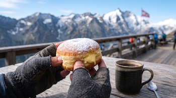 Unas manos con guantes sostienen un krapfen junto a una taza de café en una mesa de madera ante un paisaje de montañas nevadas.