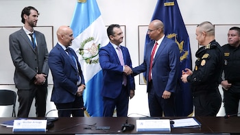 Un grupo de seis hombres en una sala de reuniones; dos hombres de traje se dan la mano frente a dos banderas de Guatemala mientras otros observan