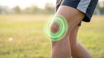 Primer plano de una rodilla masculina con pantalones cortos oscuros, mostrando un círculo verde brillante sobre la articulación en un campo verde borroso.