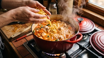 Manos vertiendo fideos cortos en una cacerola roja con caldo hirviendo, pollo y verduras sobre la hornalla de una cocina.