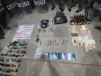 Un extenso decomiso de drogas, parafernalia, celulares, dinero, cigarrillos y alcohol sobre pavimento, con policía militar al fondo